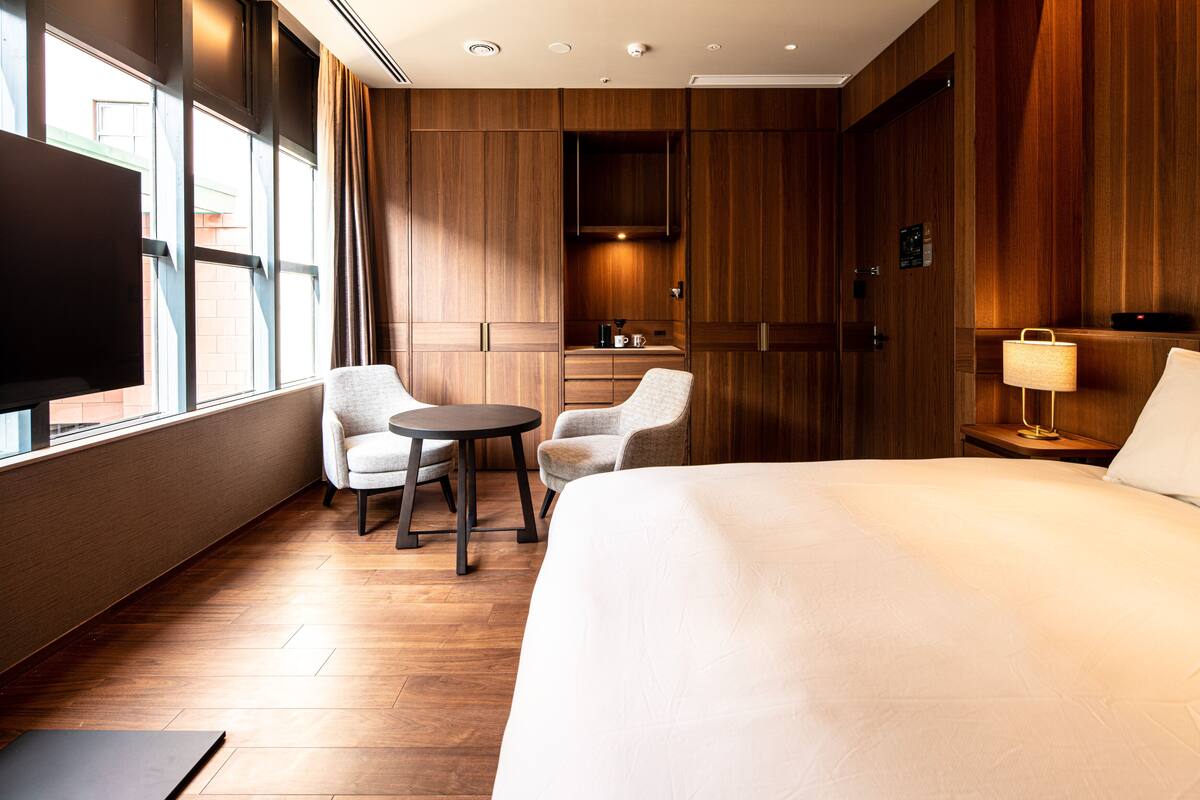 A hotel room with a large bed, two armchairs, a small table, a TV, and a wooden wardrobe.