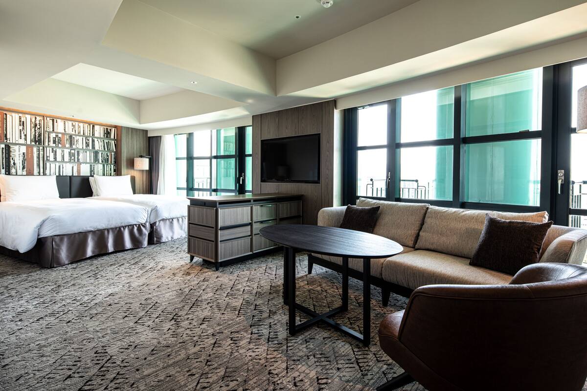 A modern hotel room with a bed, sofa, TV, and a large window.