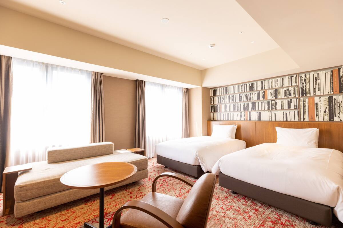 A hotel room with two beds, a sofa, a small table, and a bookshelf.