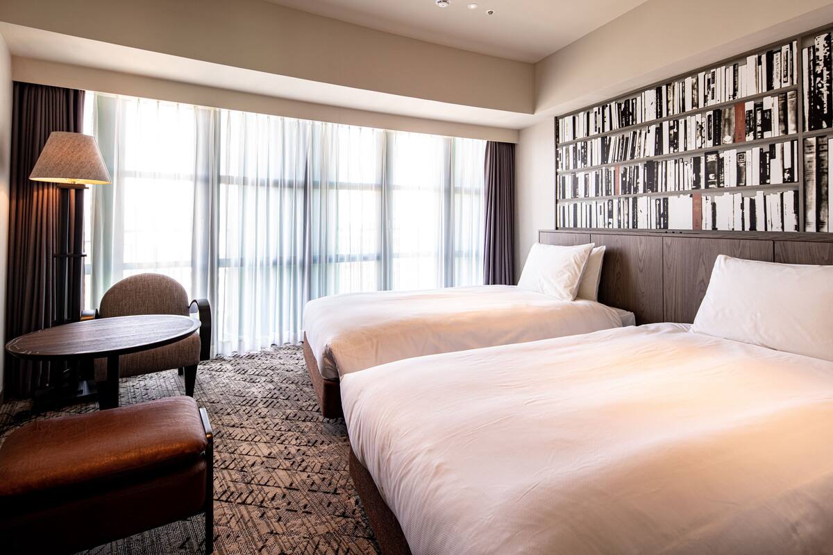 A hotel room with two beds, a small table, a chair, and a bookshelf.