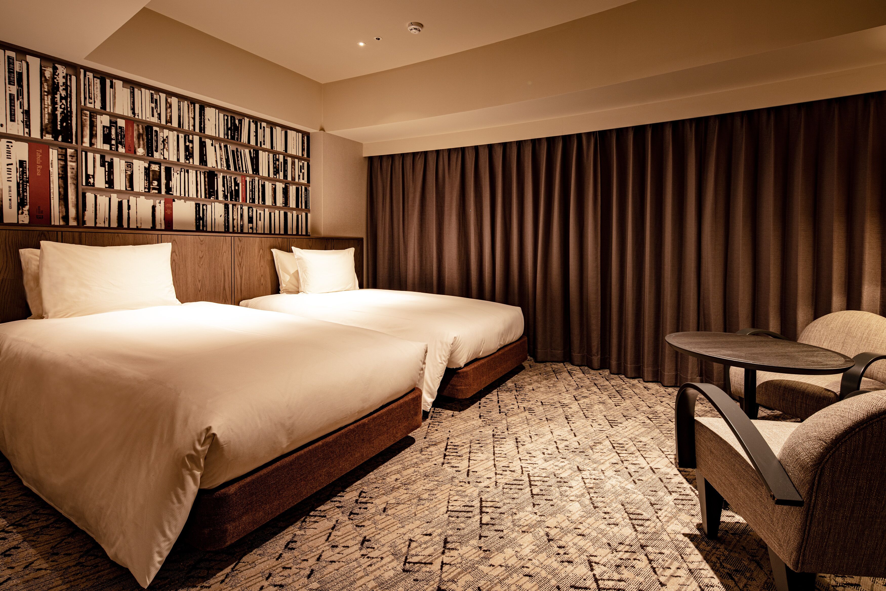 A hotel room with two beds, a bookshelf, a small table, and a large window with curtains.