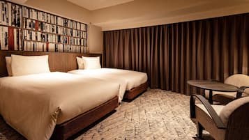 A hotel room with two beds, a bookshelf, a small table, and a large window with curtains.