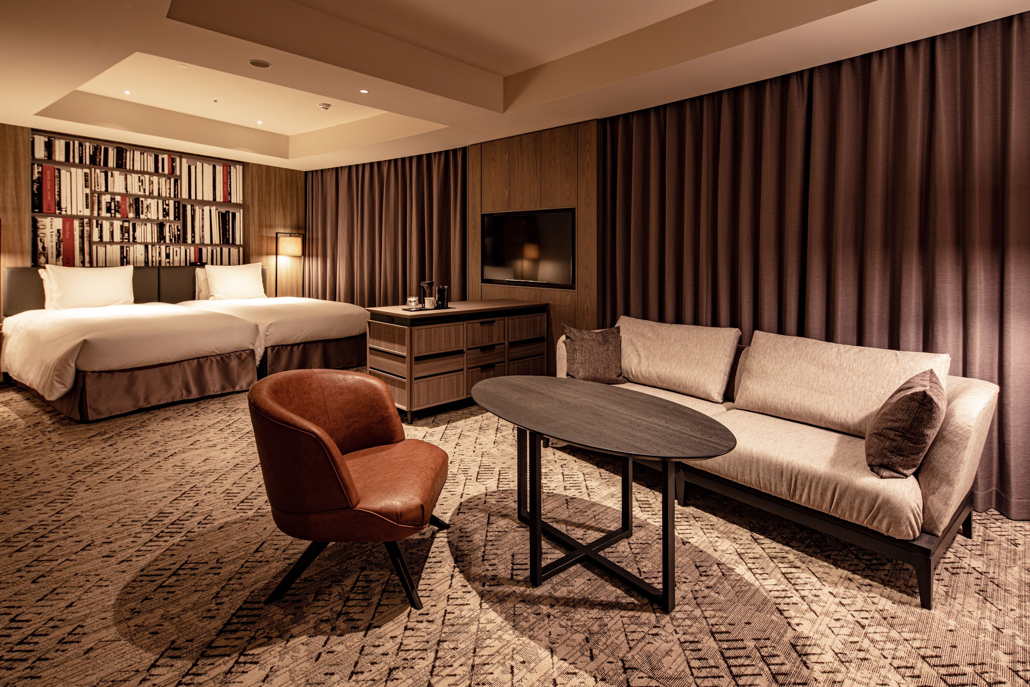 A hotel room with a bed, a sofa, a chair, a coffee table, a TV, and a bookshelf.
