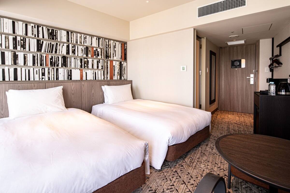 A hotel room with two beds, a bookshelf, a desk, and a dining table.