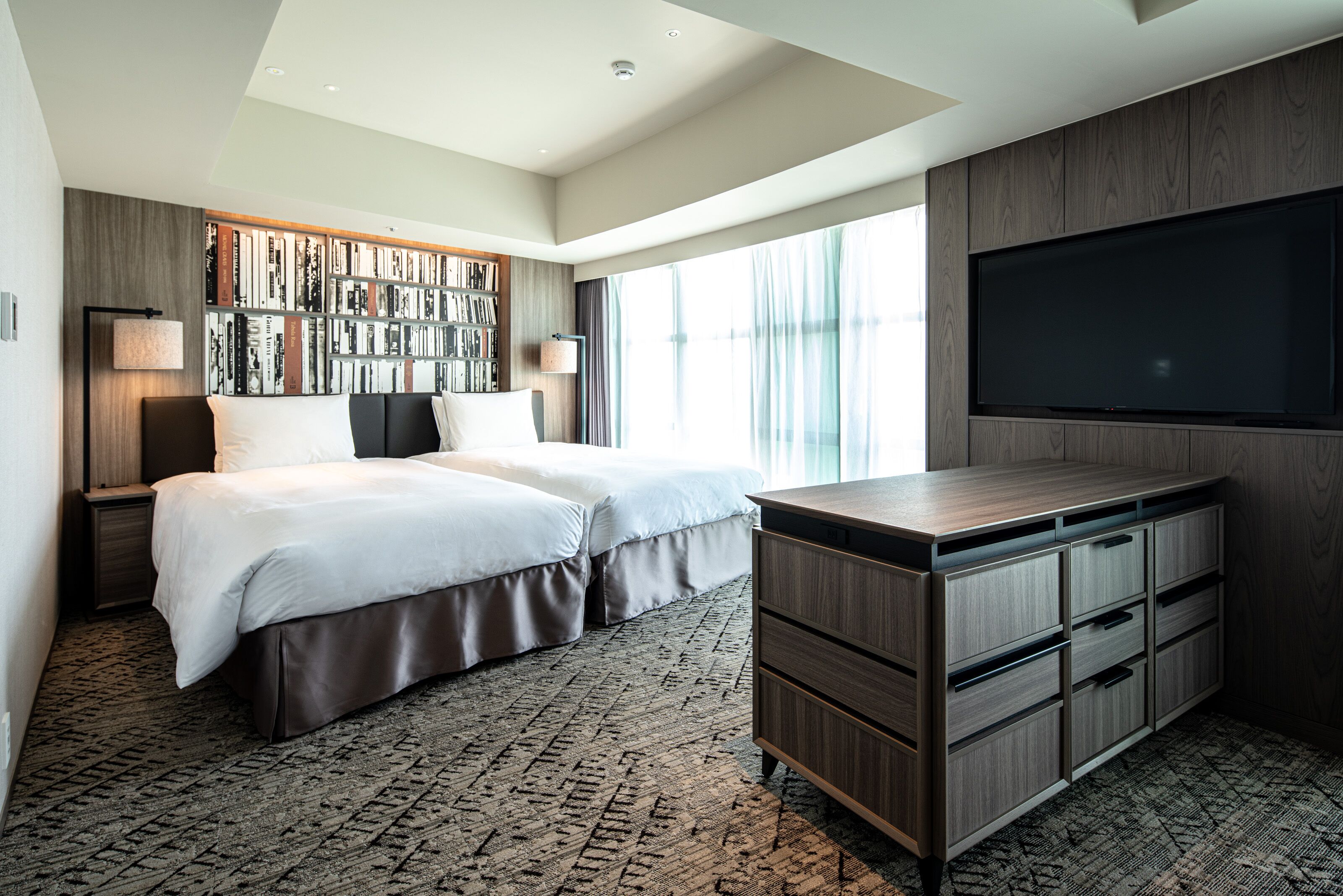 A modern hotel room with a large bed, a built-in bookshelf, a flat-screen TV, and a desk with drawers.