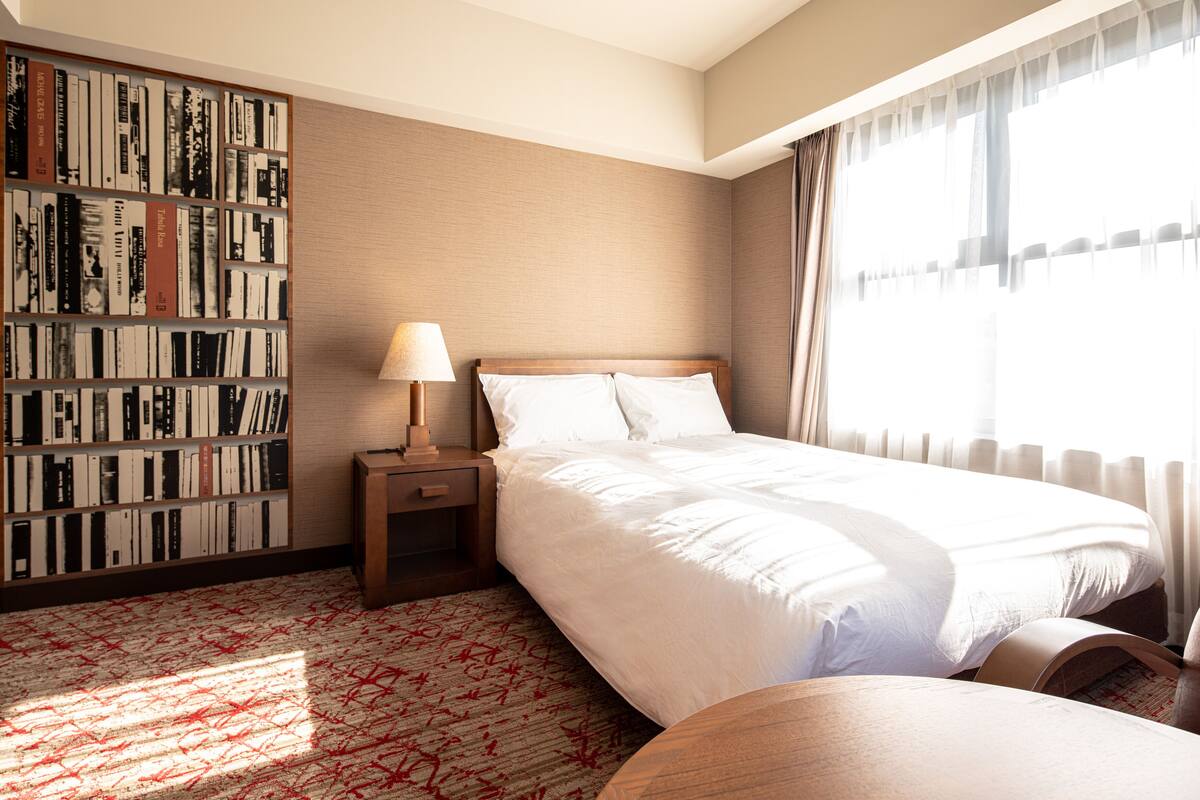 A hotel room with a large bed, a bedside table with a lamp, a bookshelf, and a large window with curtains.