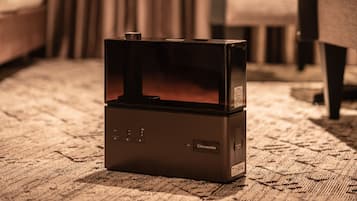 Un dispositivo electrónico negro sobre una alfombra en una habitación con una cama y cortinas al fondo.