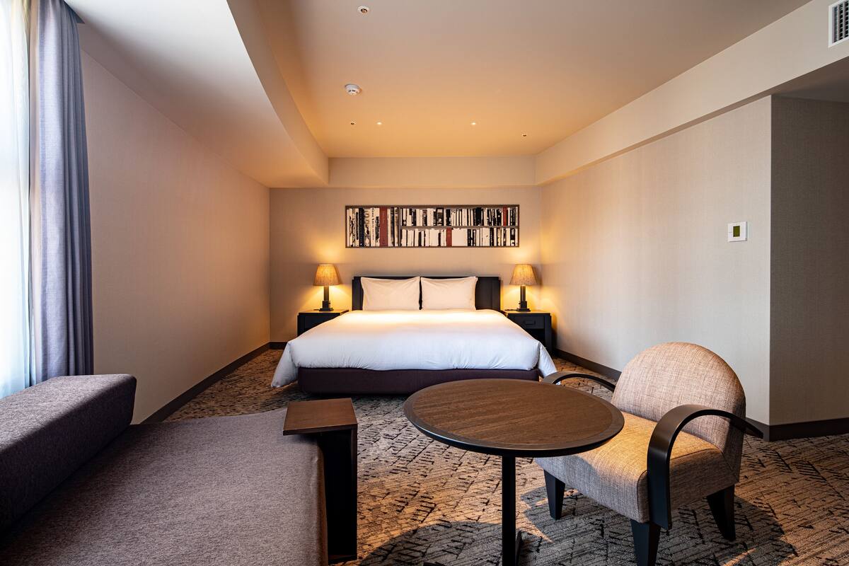 A hotel room with a bed, two bedside lamps, a round table, a chair, and a carpeted floor.