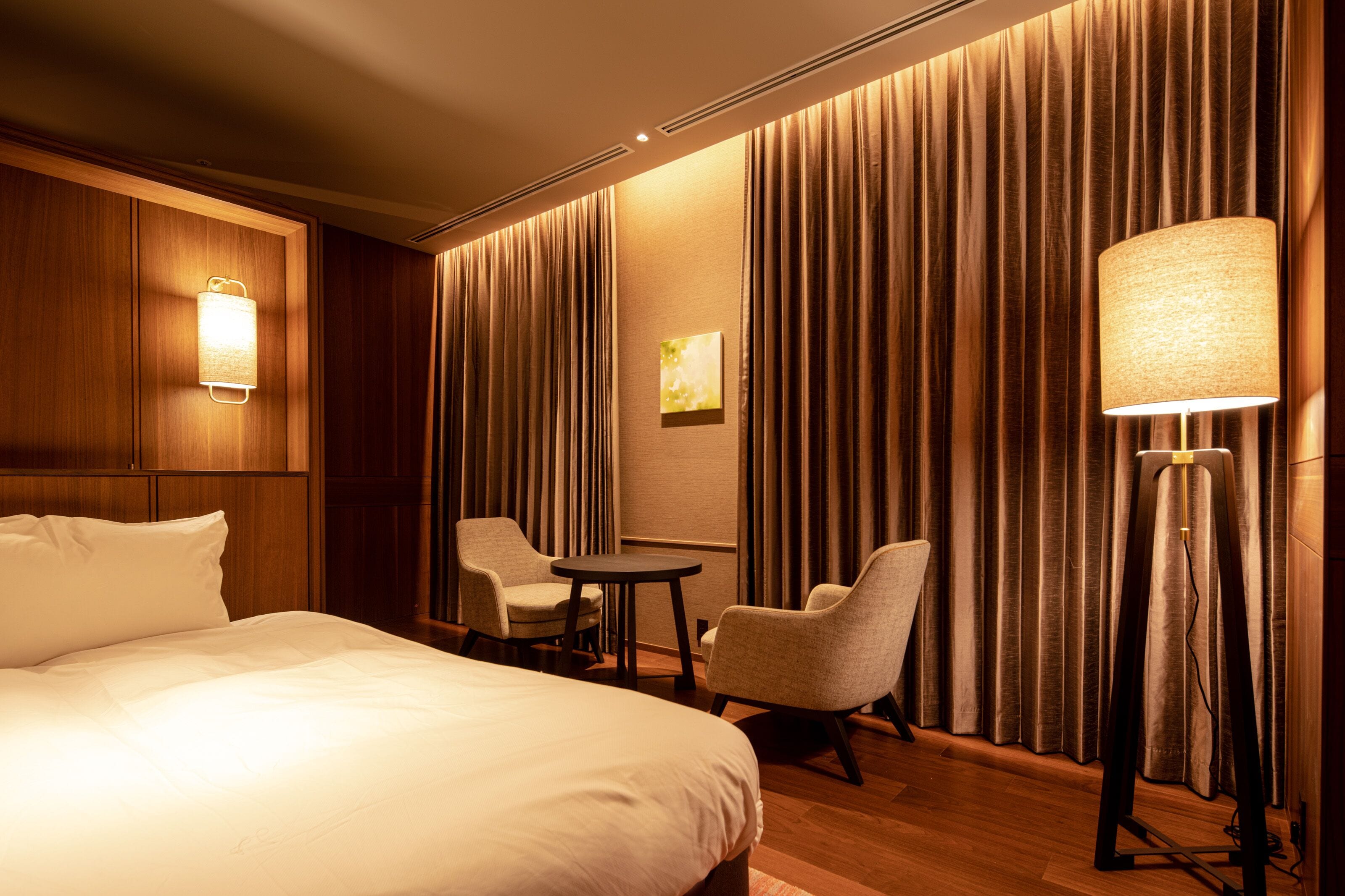 A hotel room with a large bed, two armchairs, a small table, a lamp, and a wall-mounted light fixture.