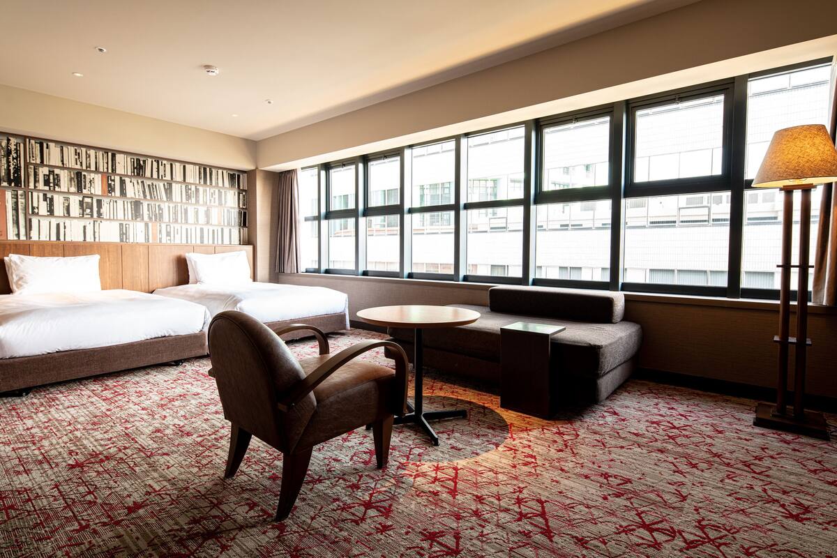 A hotel room with a large window, a bookshelf, a bed, a sofa, a chair, and a lamp.