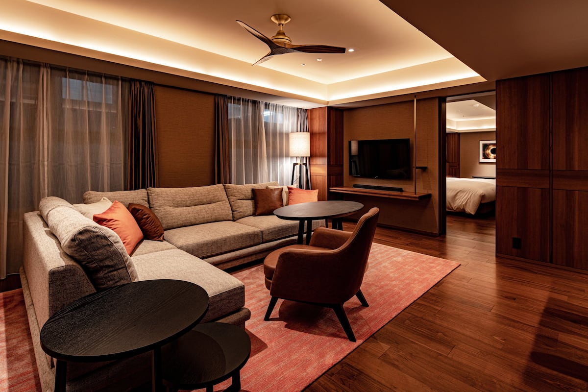 A modern hotel room with a sectional sofa, a small round table, a single armchair, a standing lamp, and a flat-screen TV mounted on the wall.