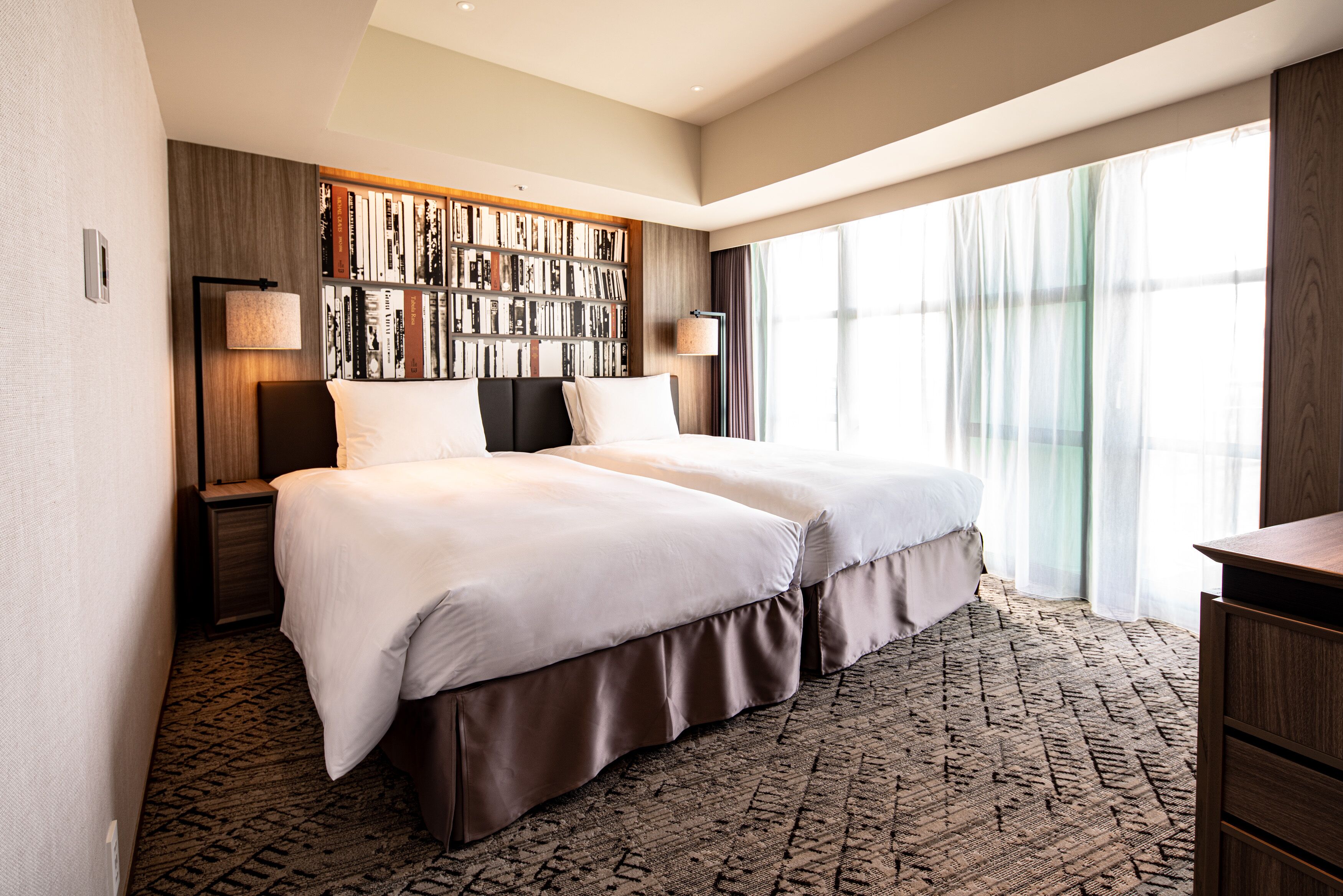 A hotel room with a large bed, a bookshelf, a desk, and a large window.