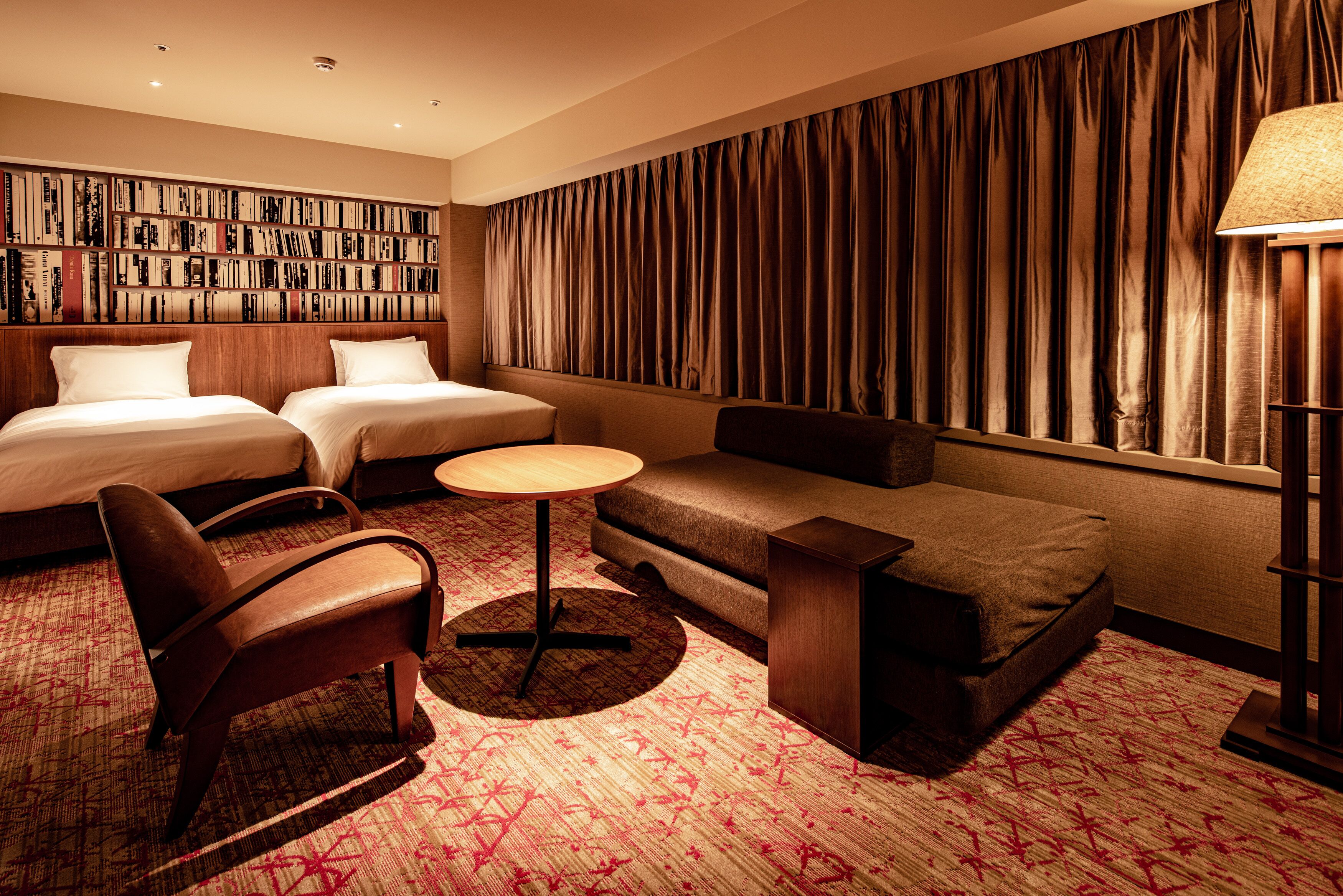 A hotel room with two beds, a sofa, a chair, a small table, a bookshelf, and a large curtain.