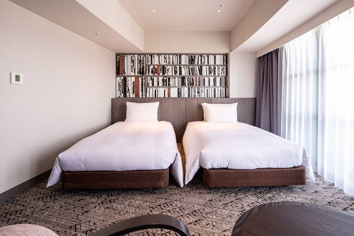 A hotel room with two beds, a bookshelf, and a large window with curtains.