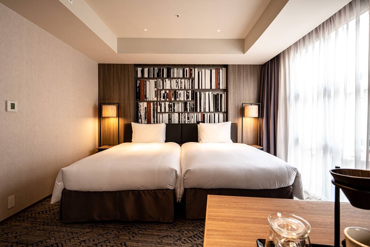 A hotel room with two beds, a bookshelf, a desk, and a large window with curtains.