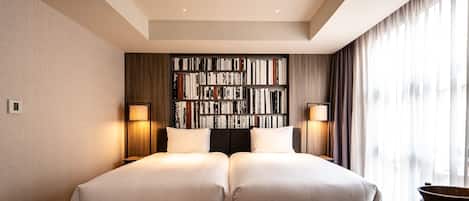 A hotel room with two beds, a bookshelf, a desk, and a large window with curtains.