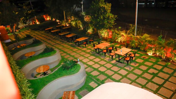 Un patio al aire libre con varias mesas y sillas dispuestas en forma semicircular, rodeado de vegetación e iluminación decorativa.