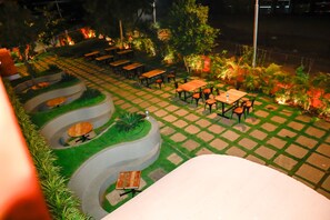 Un patio al aire libre con varias mesas y sillas dispuestas en forma semicircular, rodeado de vegetación e iluminación decorativa.