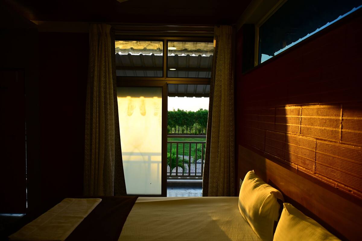 A bedroom with a bed, a window with curtains, and a view of a balcony with greenery.