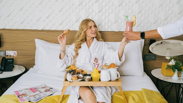 Habitación de hotel con cama, mesita de noche con teléfono, bandeja con alimentos para el desayuno y una revista sobre la cama.