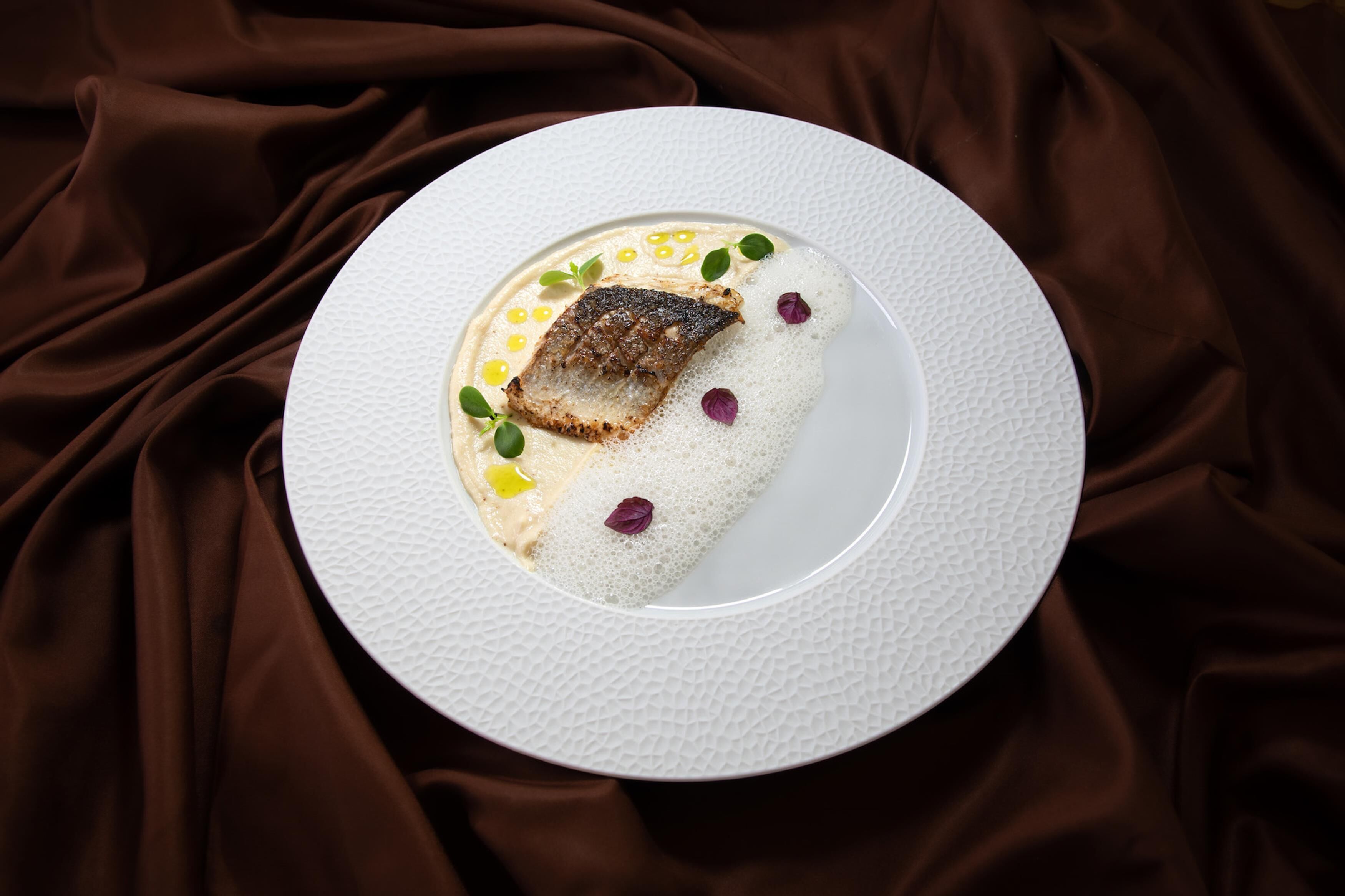 A white plate with a piece of fish and a creamy sauce, garnished with small flowers and corn kernels, on a textured white plate.