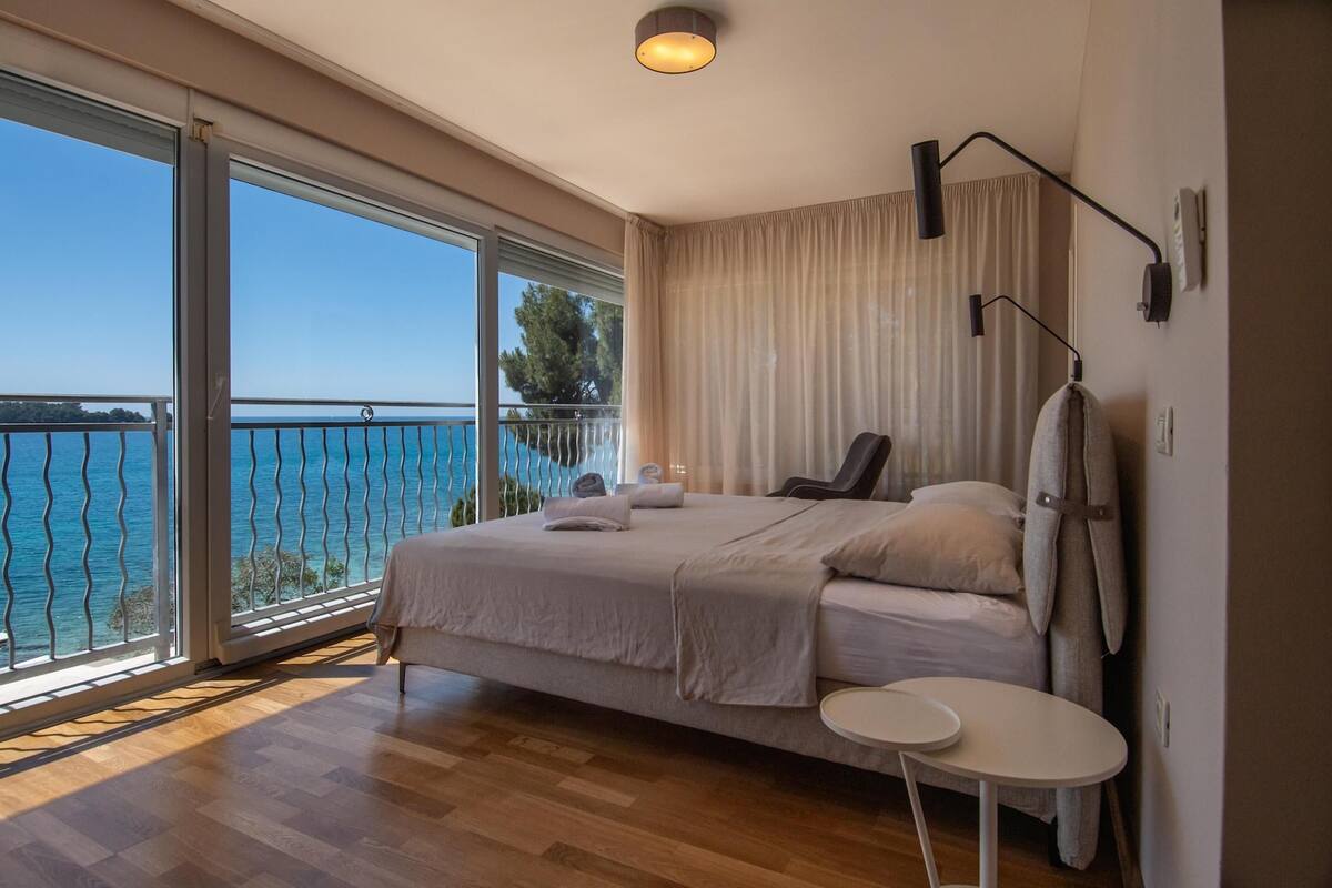 Un dormitorio con una cama grande, una mesita redonda pequeña, un balcón con barandilla y vistas al mar.