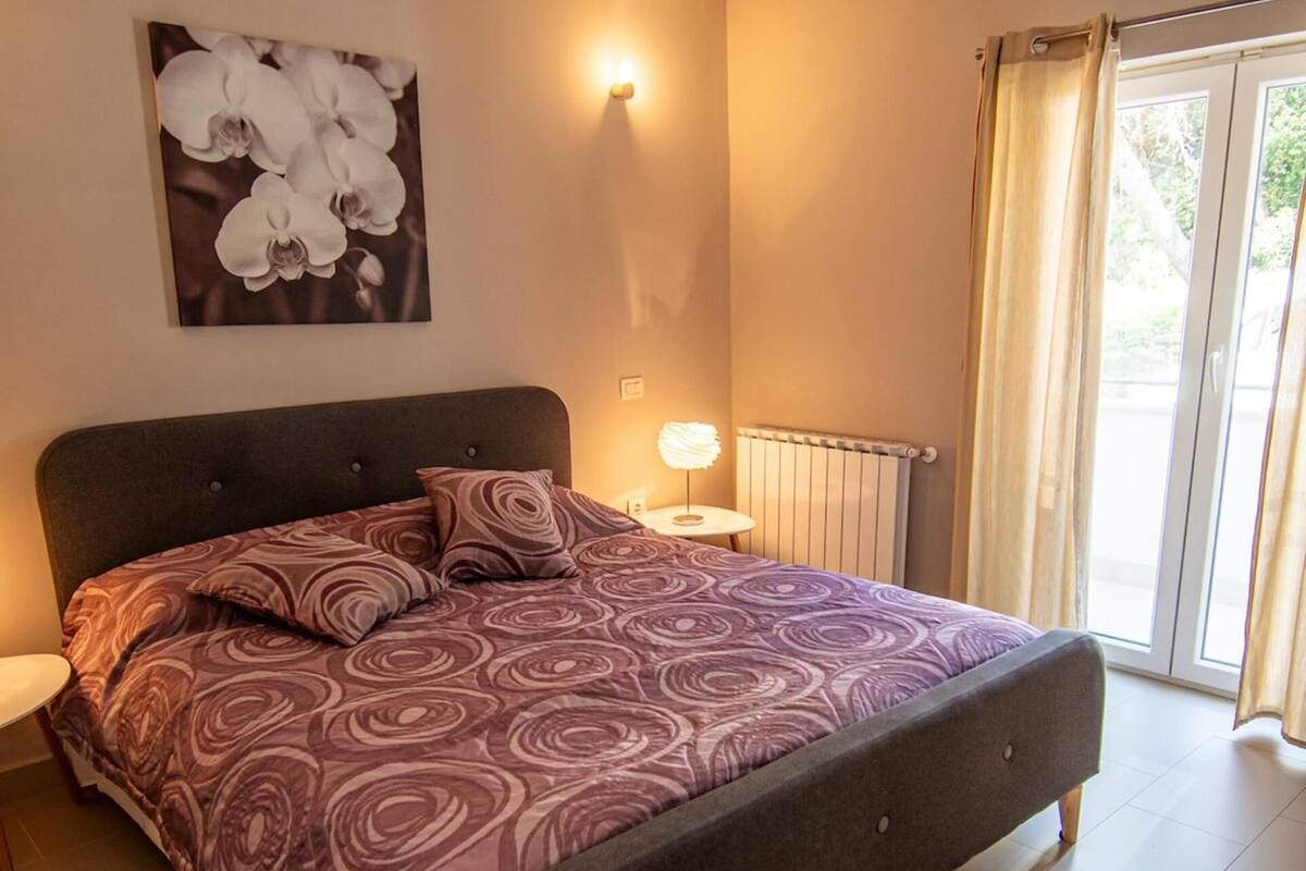 Un dormitorio con una cama grande, una obra de arte en la pared, una mesita de noche con una lámpara y vistas a través de una ventana con cortinas.