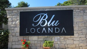 Schild für 'Blu Locanda' vor einer Steinwand, mit zwei Blumentöpfen im Vordergrund.