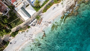 Eine Vogelperspektive zeigt einen Strand mit klarem türkisblauem Wasser, einer Holzpromenade und Gebäuden mit Balkonen.
