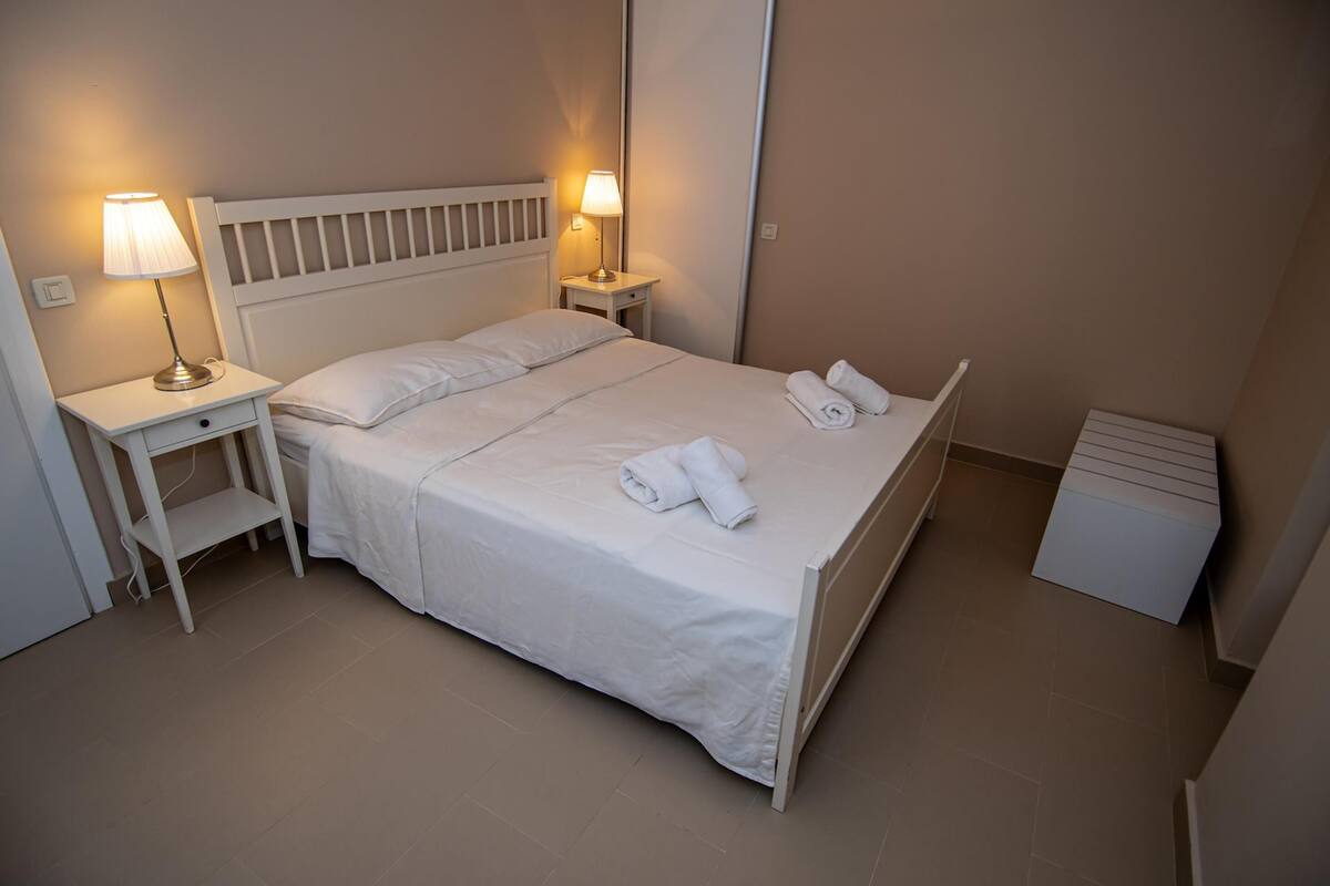 Una cama bien hecha con sábanas y toallas blancas, dos lámparas de noche, una mesita pequeña y un mueble blanco para guardar objetos.