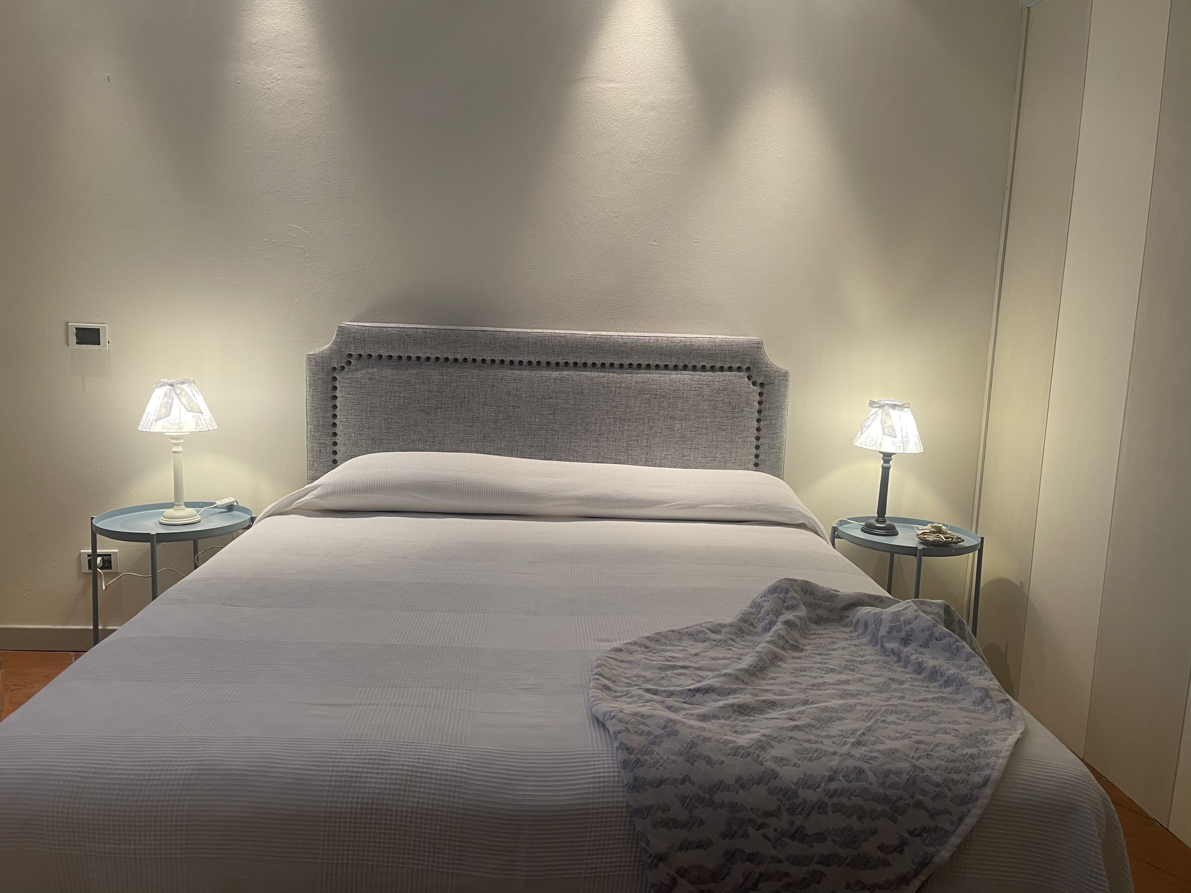 Una cama bien hecha con un cabecero tapizado gris, dos mesitas de noche con lámparas y una manta decorativa estampada.