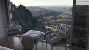 Una habitación con un ventanal que ofrece vistas a un valle boscoso. Hay dos sillas con cojines, uno con un cojín estampado y otro con un cojín floral.