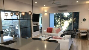 Una cocina moderna con una isla central, un comedor con mesa y sillas, una sala de estar con sofá y chimenea, y ventanas amplias que ofrecen vistas al exterior.