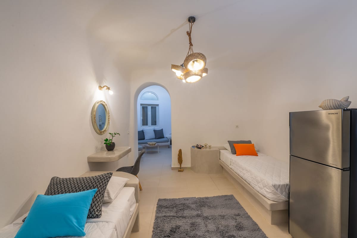 Een compacte, goed verlichte kamer met een kitchenette, een bed met een blauw kussen, een bank met gestreepte kussens en een kleine tafel met een plantje.