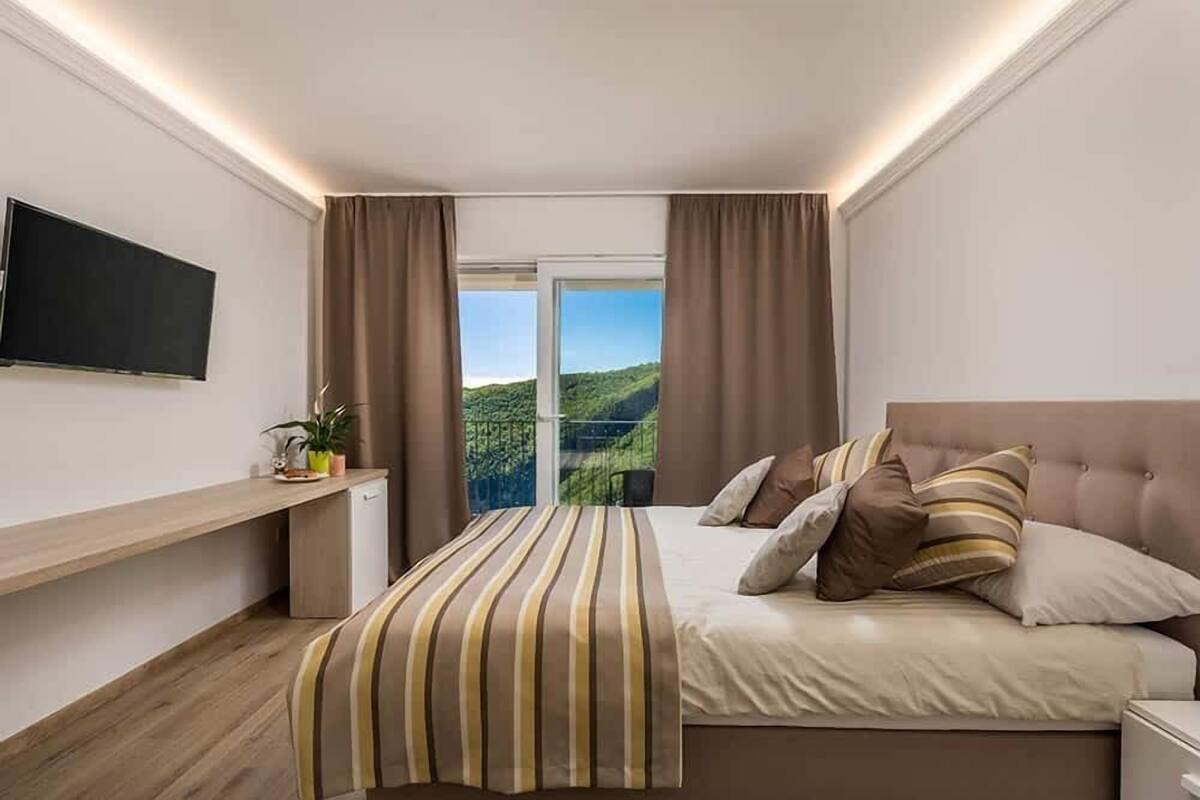 Ein modernes Schlafzimmer mit Bett, einem an der Wand befestigten Fernseher, einem Schreibtisch und Blick ins Grüne.
