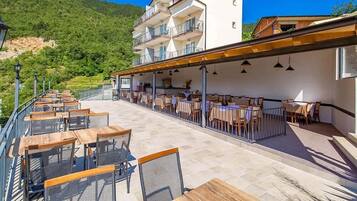 An outdoor terrace with wooden tables and chairs, a covered seating area, and a view of a hillside.