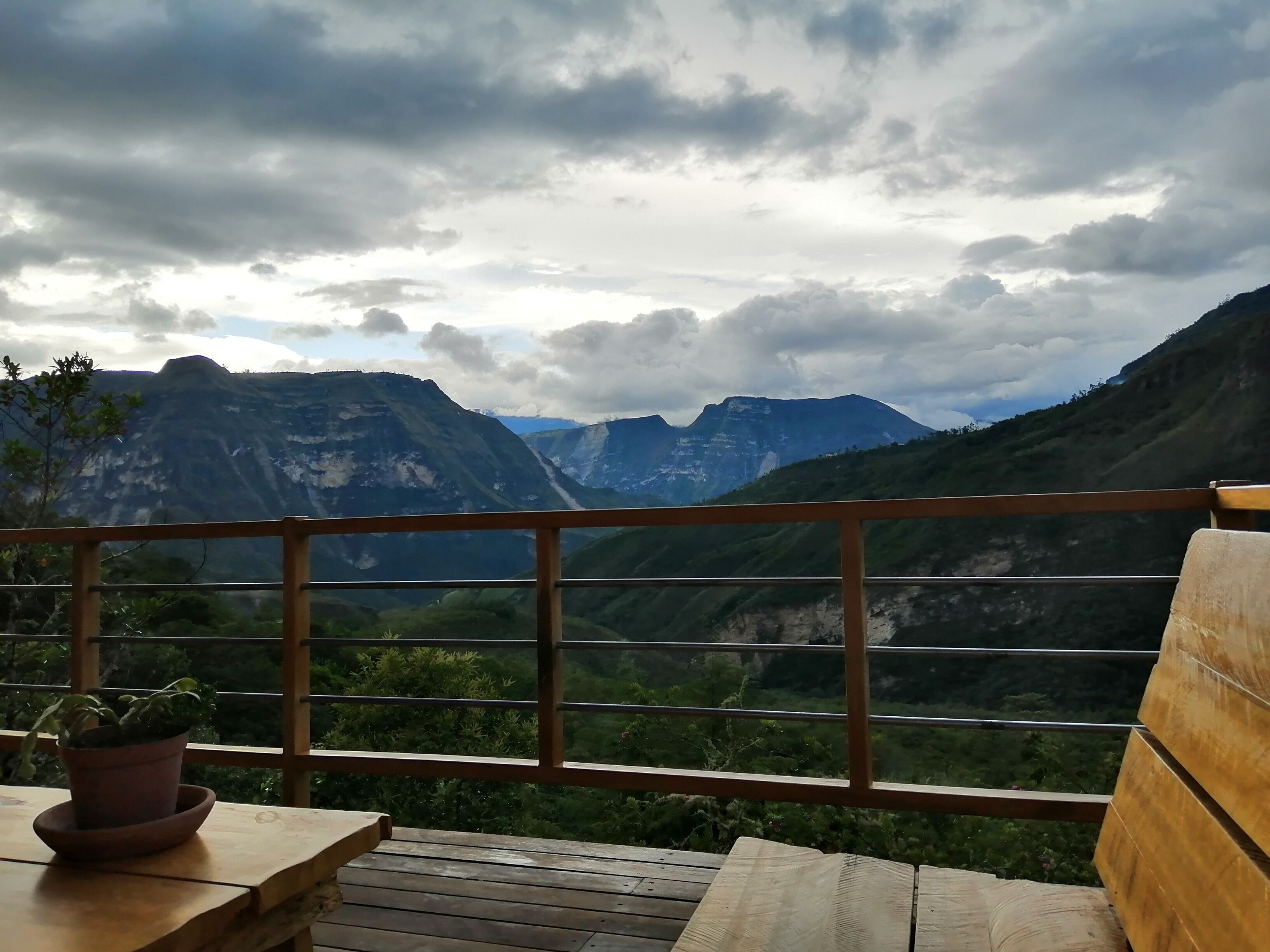 A wooden deck with a potted plant, overlooking a mountainous landscape with a cloudy sky.