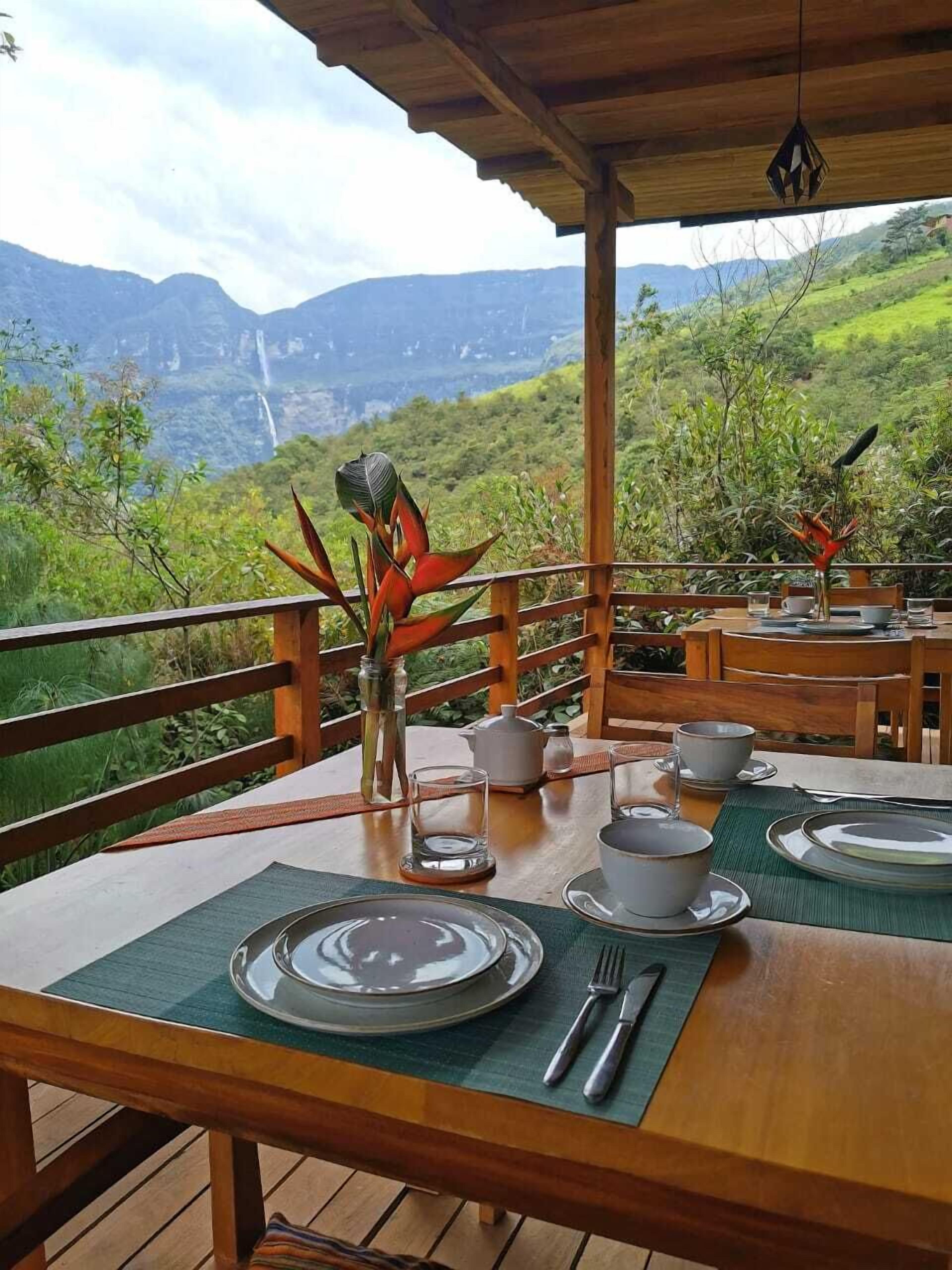 Mesa posta com pratos, tigelas e talheres, rodeada por cadeiras de madeira, com vista para vegetação exuberante e montanhas ao fundo.