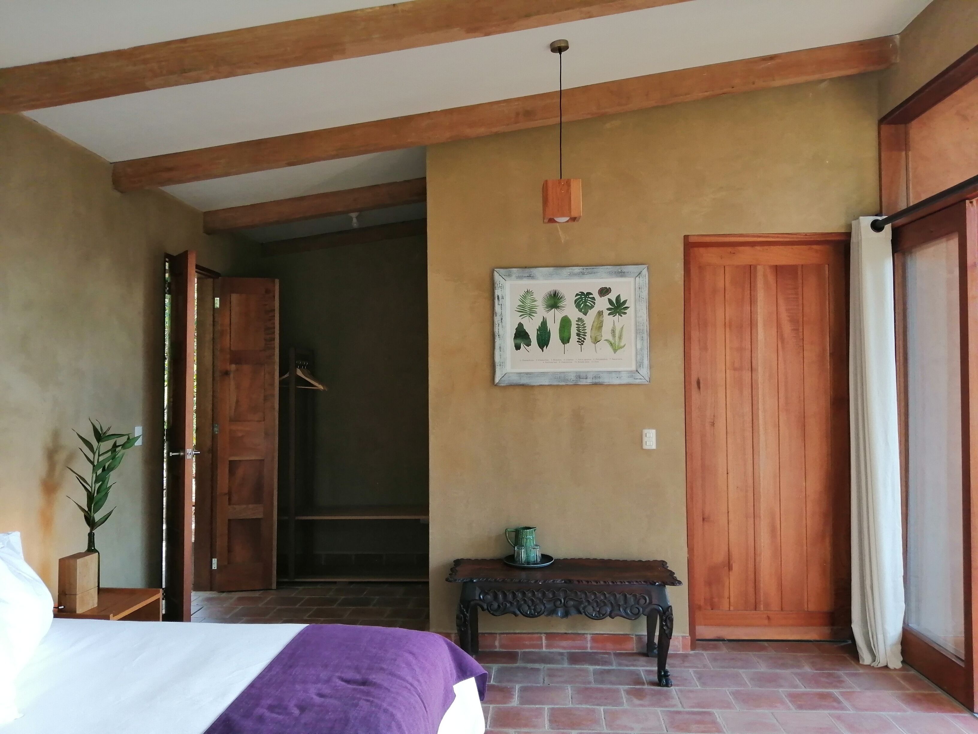 Un dormitorio con una cama, un banco de madera, una planta montada en la pared, una impresión botánica enmarcadas y una puerta de madera.
