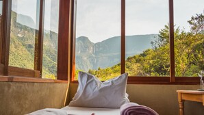 A room with a bed, pillows, and towels, with a view of green hills and a waterfall through large windows.
