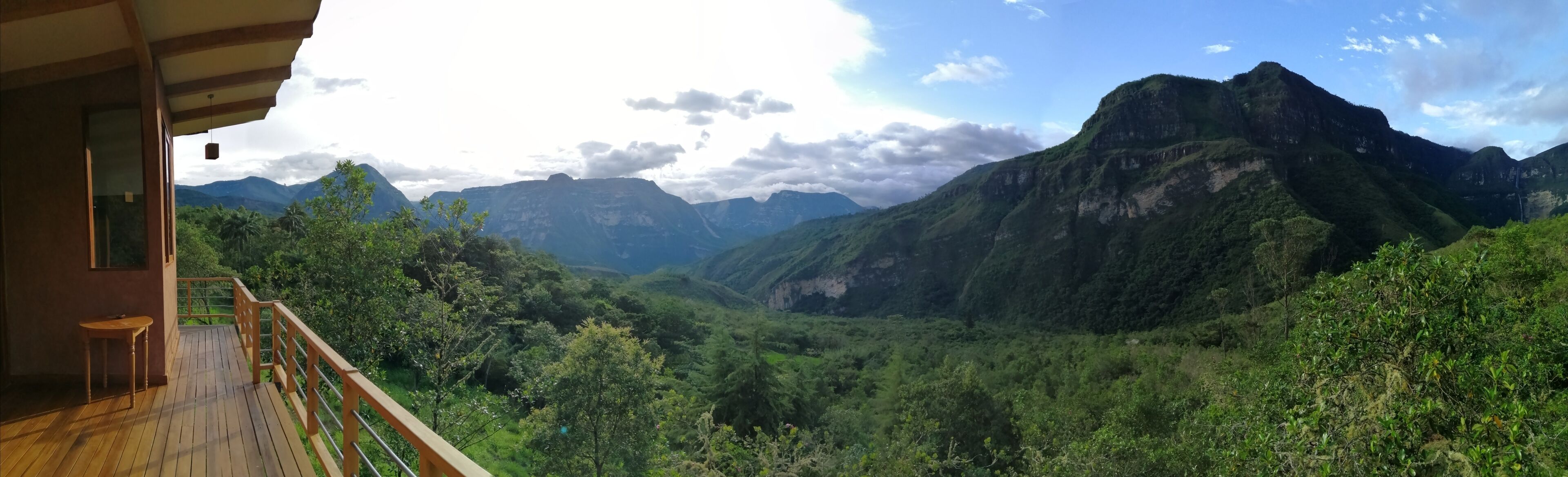Una terraza de madera con una mesa, con vista a un valle exuberante y montañas al fondo.