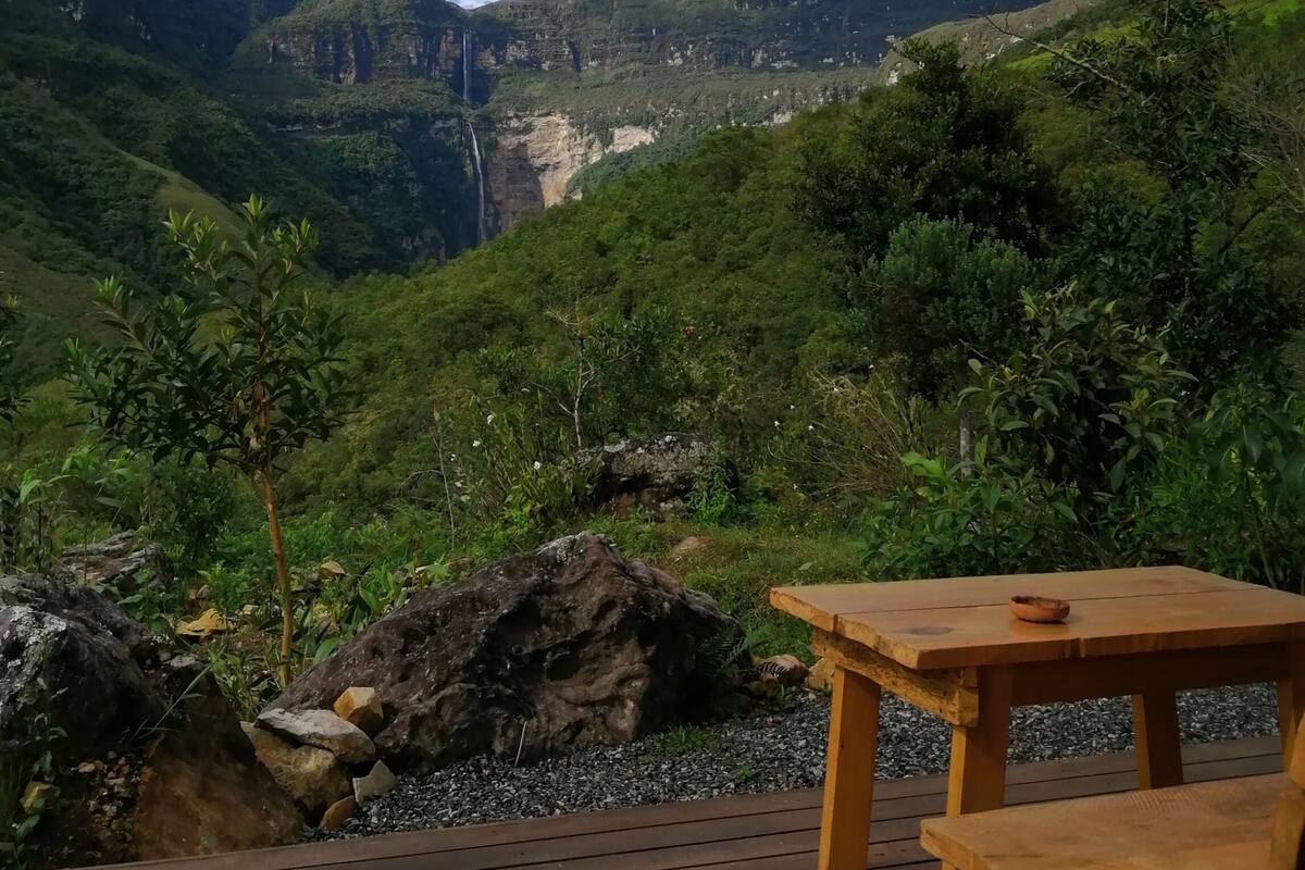 A wooden deck with a table and bench, overlooking a lush valley with a waterfall and mountains.