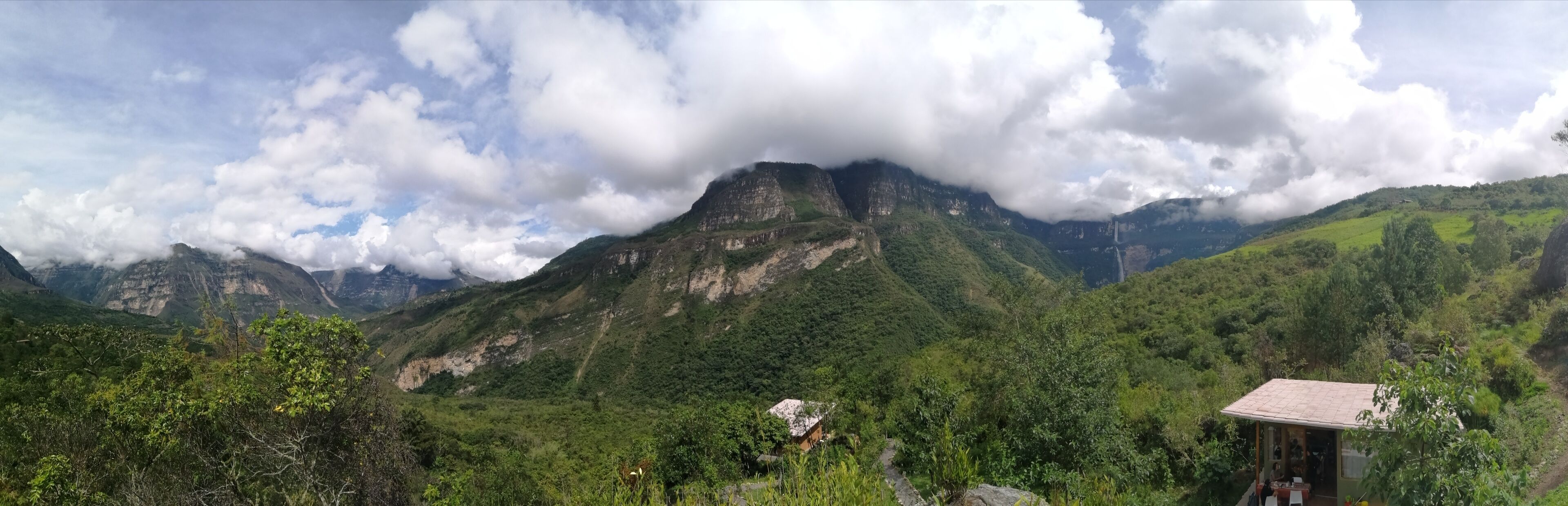 Un paisaje montañoso con una pequeña estructura de madera, posiblemente una cabaña o choza, rodeada de vegetación exuberante y un pico de montaña destacado al fondo.