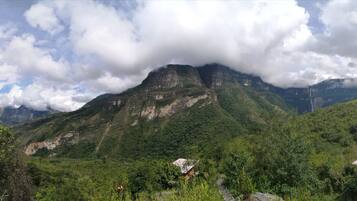 Un paisaje montañoso con una pequeña estructura de madera, posiblemente una cabaña o choza, rodeada de vegetación exuberante y un pico de montaña destacado al fondo.
