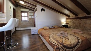 A bedroom with a large bed, a desk, two white chairs, a stone wall, and a wooden ceiling. - Studio Isabel (Rovinj)