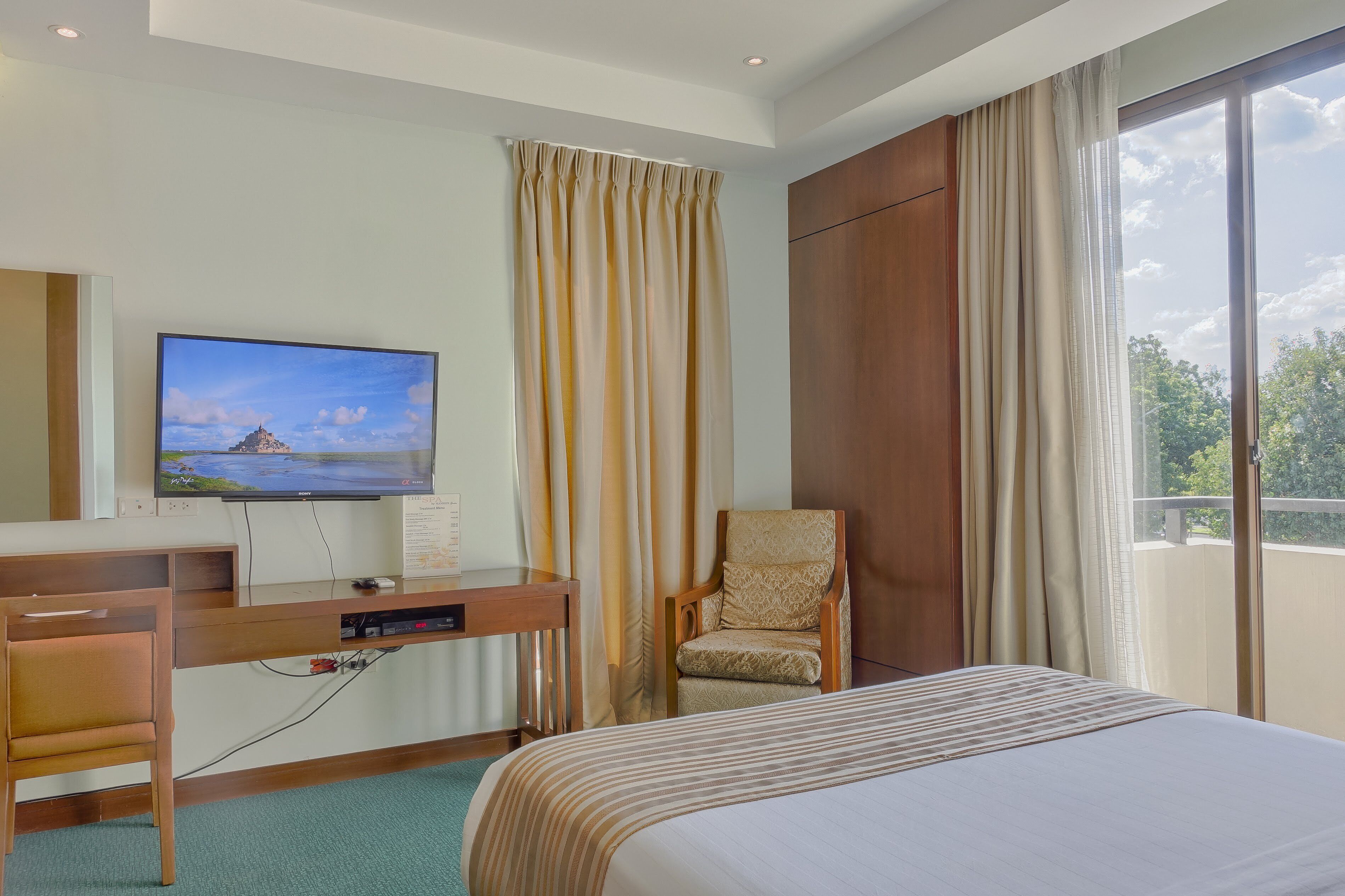 A hotel room with a bed, a desk with a television, a chair, and a large window with curtains.