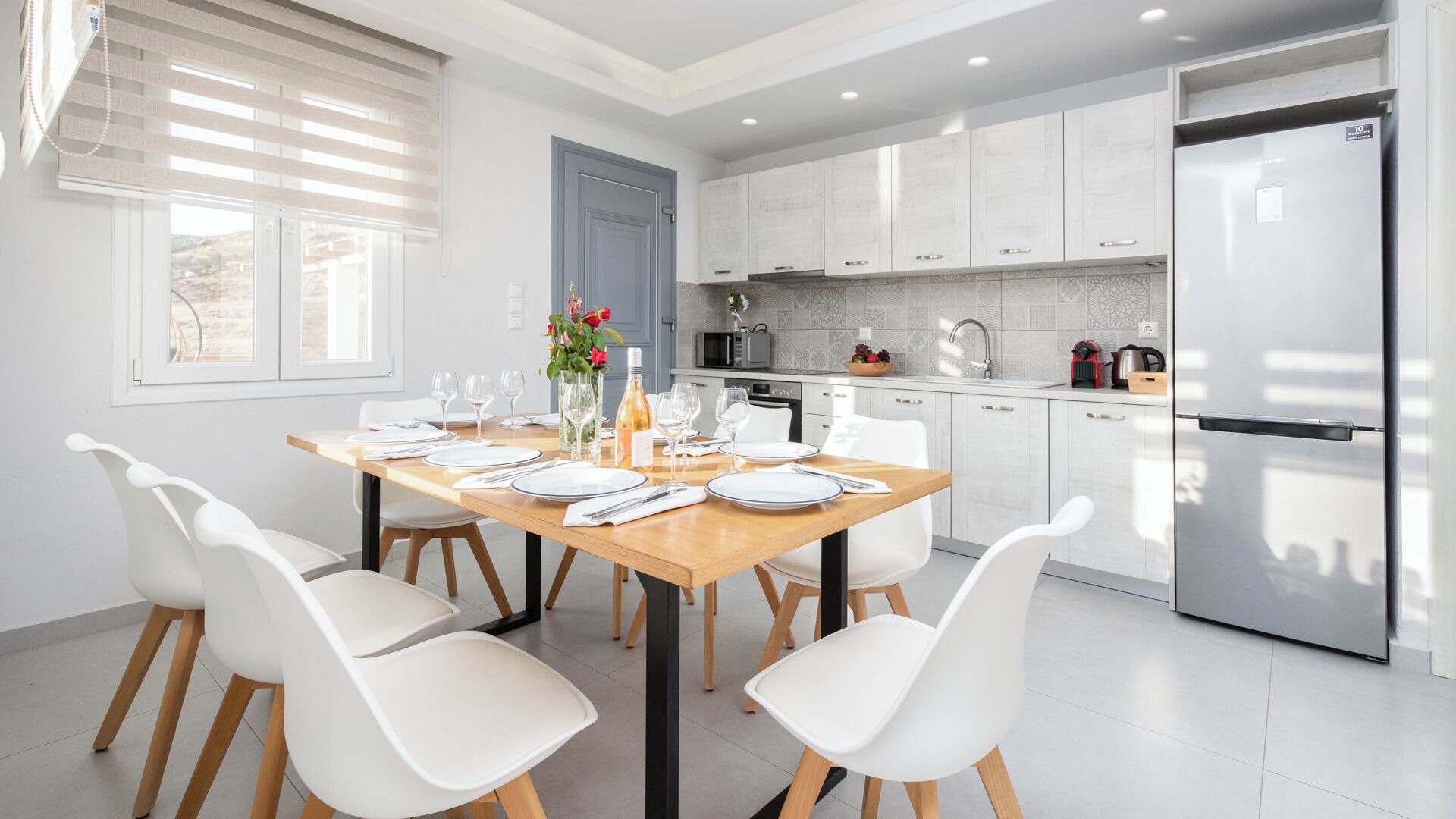 A modern kitchen with white cabinets, a stainless steel refrigerator, and a dining area with a wooden table and white chairs.
