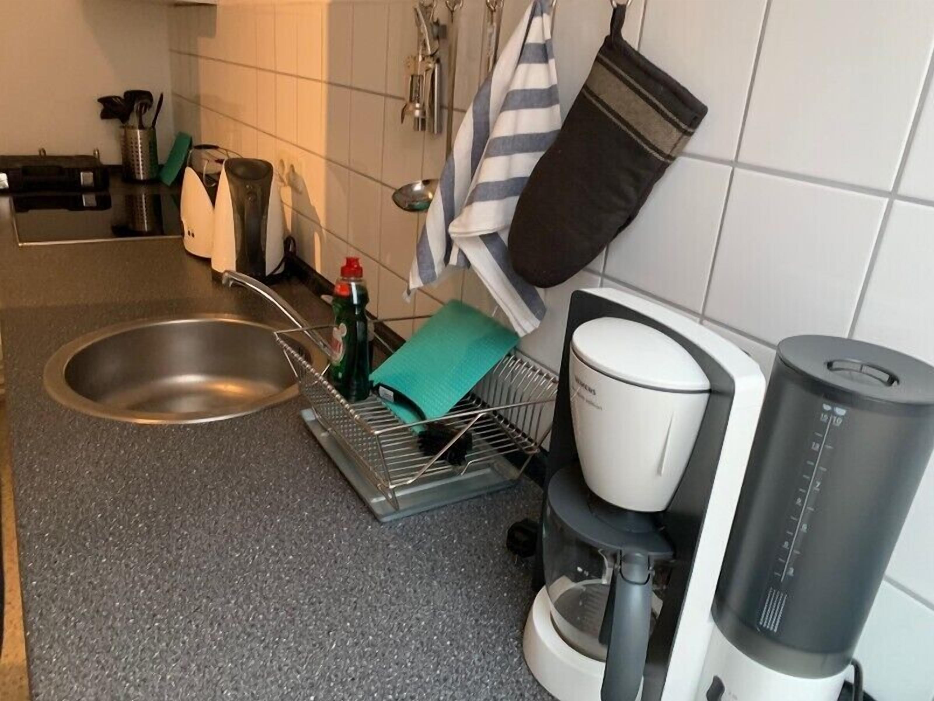 Una cocina con un lavamanos de acero inoxidable, un escurreplatos, una cafetera y un cubo de basura.