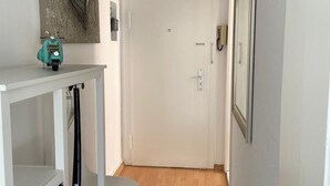 Ein schmaler Flur mit einer weißen Tür, einem kleinen Tisch mit einem Rollstuhl, einem farbenfrohen Teppich und einem gerahmten Bild an der Wand.