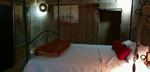Een eenpersoonsbed met een wit laken en een rood kussen, een kleine tafel met een rode tafelkleed, en een wandlamp.